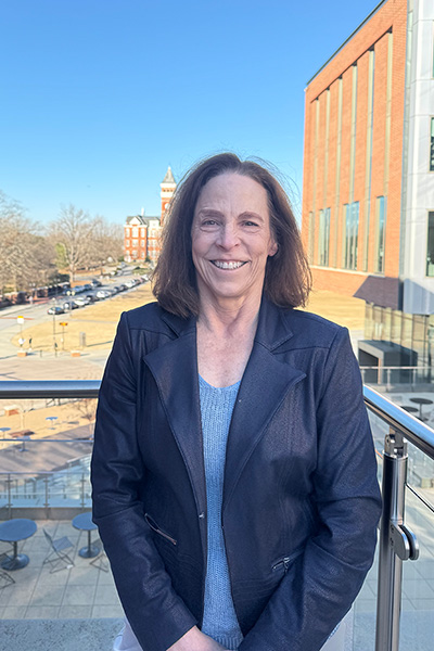 Professional portrait of a woman with shoulder-length brown hair, standing outdoors with a university campus backdrop.