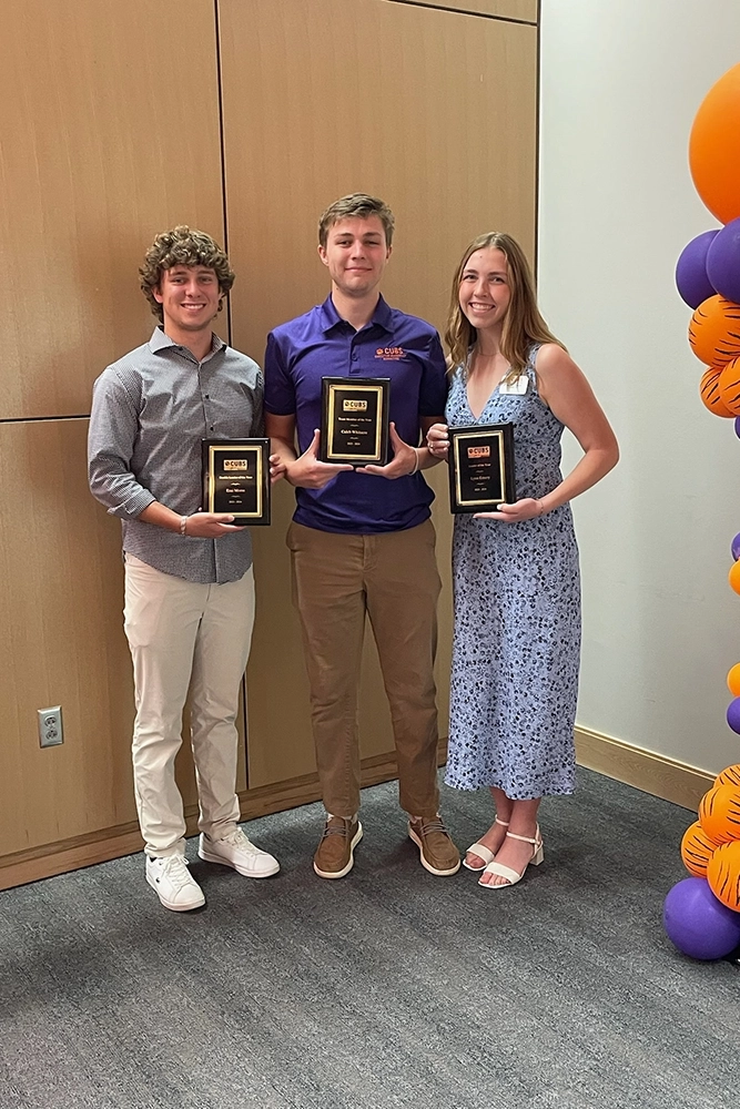 Three smiling students holding award plaques, standing together at a ceremony decorated with orange and purple balloons.