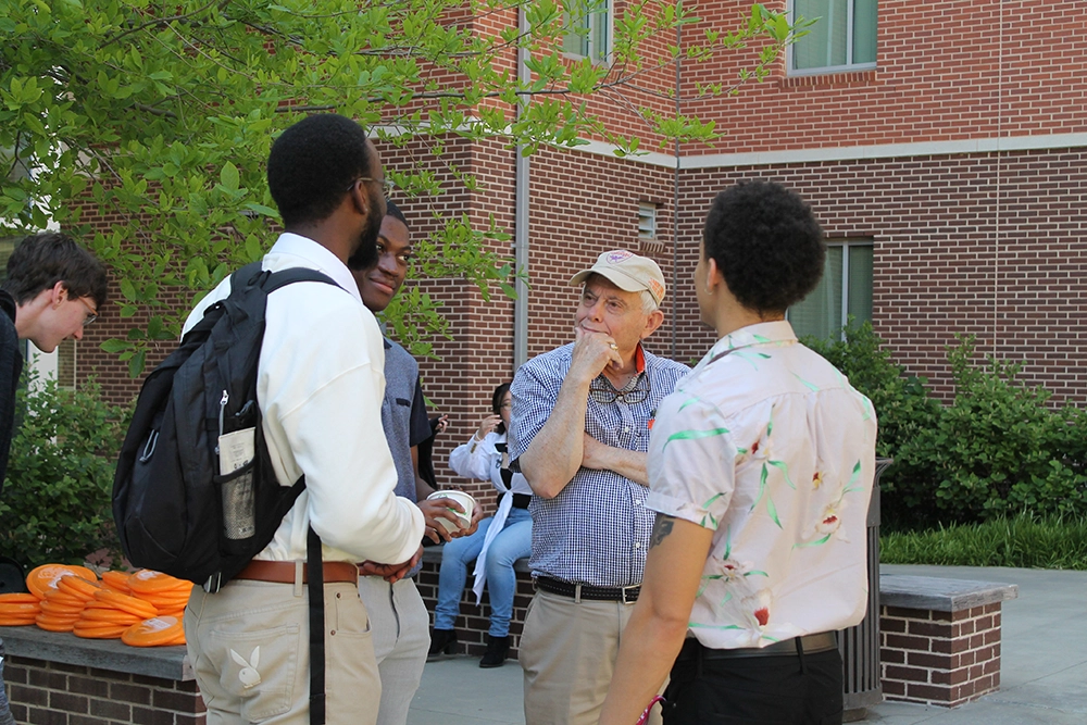 A group of diverse individuals engaged in conversation outdoors, with a focus on an older man in a cap speaking to three younger men.