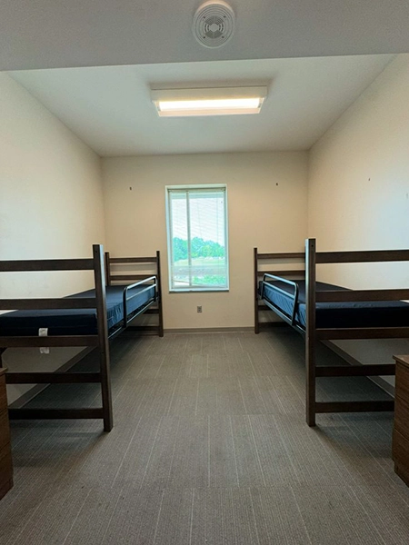 A simple dorm room featuring two unmade bunk beds and a window with a view of greenery outside.