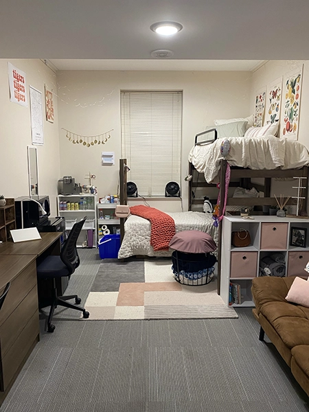 A cozy college dorm room featuring a lofted bed, a study area with a desk and chair, and personal decorations on the walls. The space is organized with storage units and has a warm, inviting atmosphere.