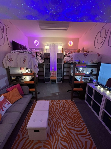 A cozy college dorm room featuring two lofted beds, a bright orange rug, and stylish lighting that creates a warm atmosphere.