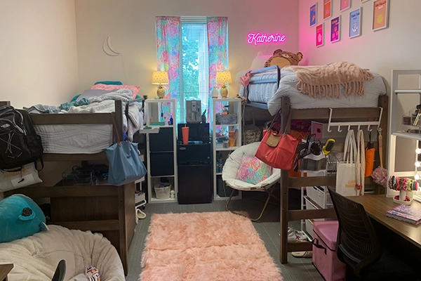 A vibrant college dorm room featuring two elevated beds, decorative string lights, and personalized decorations, including a neon sign. The room is filled with colorful furnishings and cozy accessories.