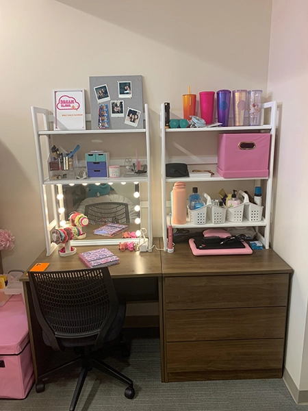 A well-organized study desk setup featuring a mirror with lights, shelves holding colorful cups and office supplies, and a chair, ideal for a college dorm.