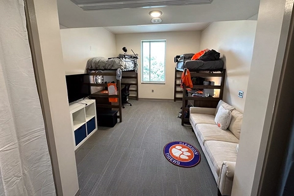 A dorm room featuring two bunk beds, a TV unit, and a couch, with Clemson Tigers branding on a rug.