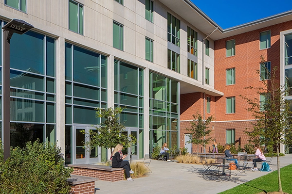 Modern college building with large glass windows and outdoor seating areas. Students are seen socializing and relaxing in the sunny courtyard.