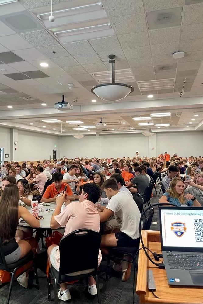 A crowded auditorium filled with students sitting at tables during an opening meeting for a club.