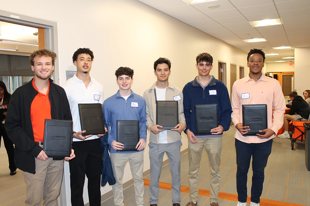 Group of six young men holding folders while posing in a corridor, likely at an academic or professional event.