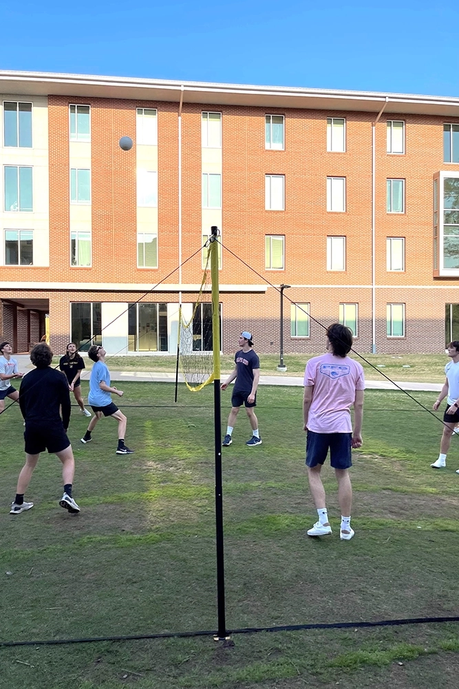 Students playing volleyball on a grassy area in front of a brick building.