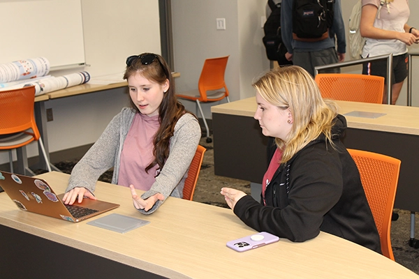 Two students engaged in a discussion while working on a laptop in a classroom setting.