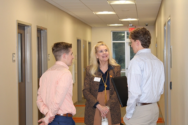 A woman is engaged in conversation with two young men in a hallway, smiling and holding a folder.