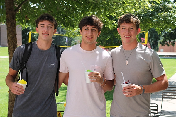 Three young men standing together outdoors, each holding a cup of colorful frozen treats in a sunny setting.