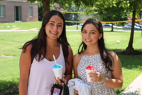 Two young women smiling while holding cups of colorful shaved ice outdoors on a sunny day.