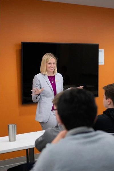 A professional woman in a light gray suit engages with an audience in a classroom setting.