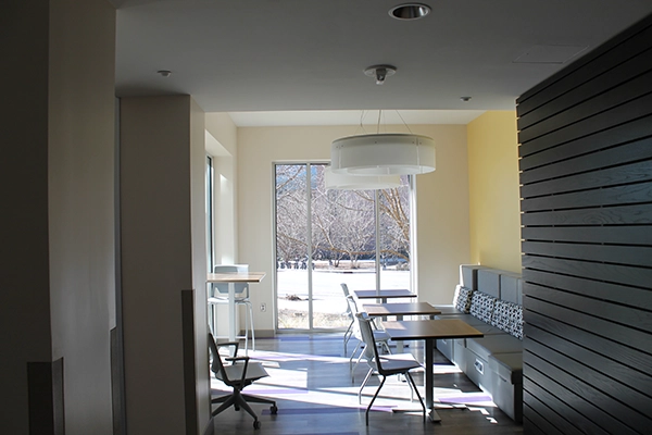 Modern coworking space featuring a bright, airy design with multiple tables and seating areas. Large windows provide natural light and a view of the outdoors.