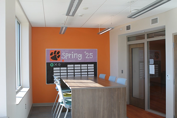 Modern conference room featuring a large table, colorful orange wall, and a display board with "Spring '25" title.
