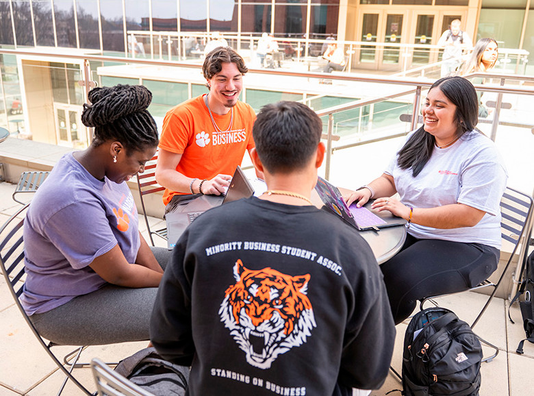 A group of diverse college students collaborating at a table outdoors while working on laptops.