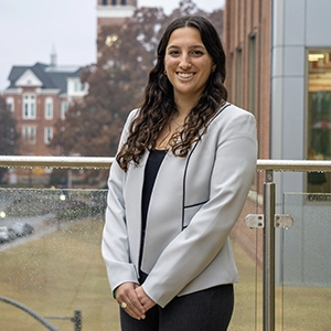 A smiling woman in a light gray blazer stands outdoors in front of a blurred university campus background.