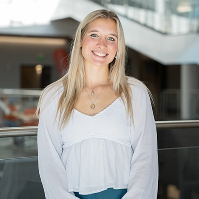 Smiling young woman with long blonde hair wearing a white blouse stands confidently in a modern atrium.