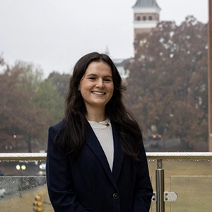 A smiling woman in a navy blazer stands outdoors with a blurred background of trees and a tower.