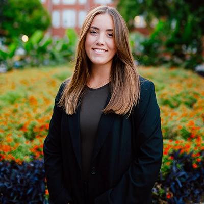 A young woman in a black blazer stands confidently against a backdrop of colorful flowers.