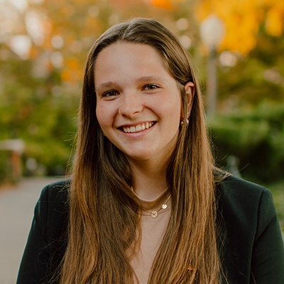 Smiling young woman with long hair wearing a blazer, surrounded by fall foliage in the background.