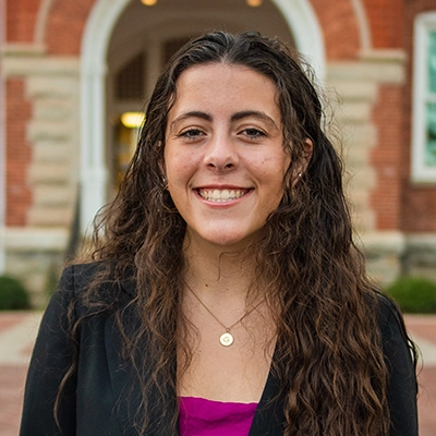 A smiling young woman with long, wavy hair, wearing a black blazer over a purple top, poses outdoors in front of a brick building.