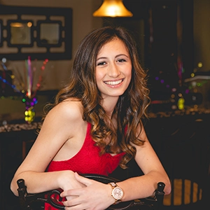 A young woman smiling while sitting at a table, wearing a red dress and a watch.