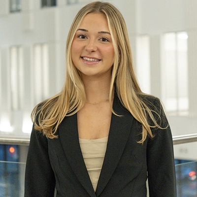 A professional headshot of a smiling young woman in a black blazer, standing indoors.