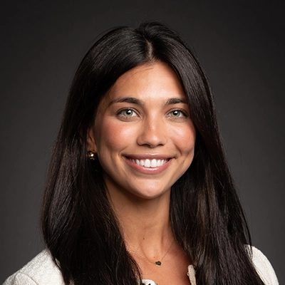 A professional headshot of a smiling woman with long dark hair and green eyes, wearing a light-colored top.