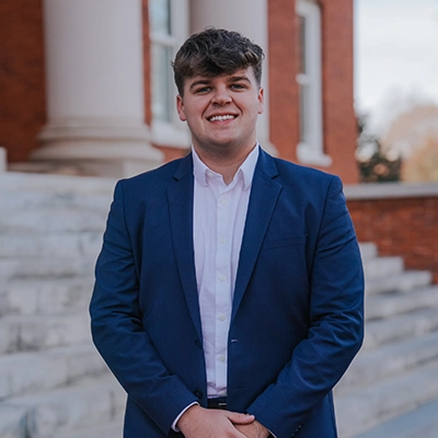 A smiling young man in a blue suit and white shirt stands confidently on a staircase in front of a brick building.