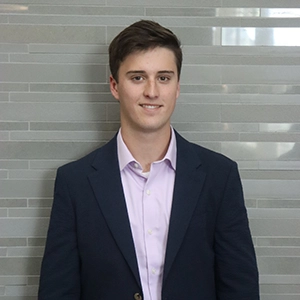 A young man wearing a blazer over a light purple shirt stands in front of a gray tiled background.