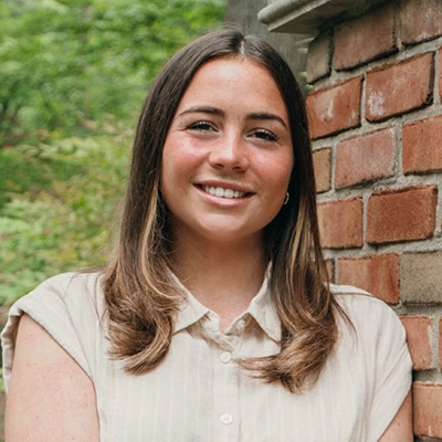 A smiling young woman stands against a brick wall, surrounded by greenery.