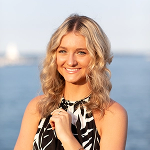 A smiling young woman with wavy blonde hair, wearing a black and white patterned top, poses against a scenic waterfront background.