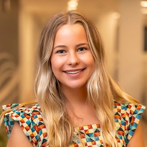 A smiling young woman with long blonde hair wearing a colorful patterned top.