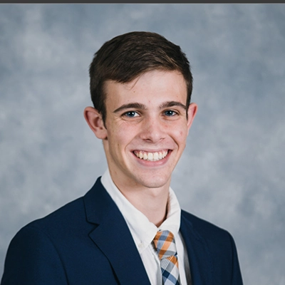 A smiling young man in a formal suit with a tie, set against a gray background.