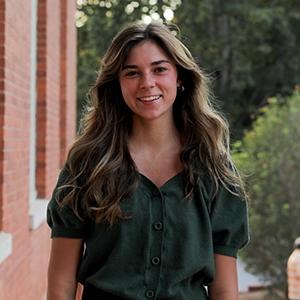 A smiling young woman with long, wavy hair wearing a dark green button-up shirt, standing outside near brick architecture.