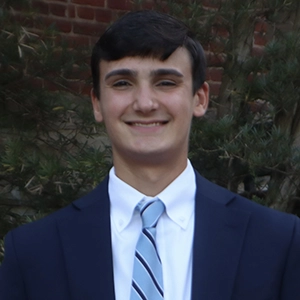 Young man in formal attire smiling outdoors, with greenery in the background.