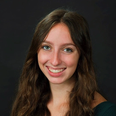 A smiling young woman with long, wavy hair and green eyes poses against a dark background.