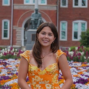 A smiling young woman in a vibrant yellow floral dress stands in a garden filled with colorful flowers, with a historic building and statue in the background.