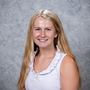 A professional headshot of a young woman with long blonde hair, wearing a white sleeveless top with a ruffled neckline, against a textured gray background.