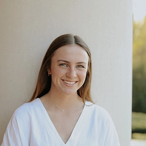 A smiling young woman in a white top, standing against a soft background.