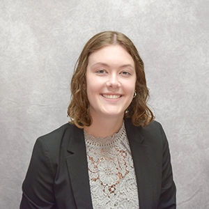 Professional headshot of a young woman wearing a black blazer and a lace top, smiling against a neutral gray background.