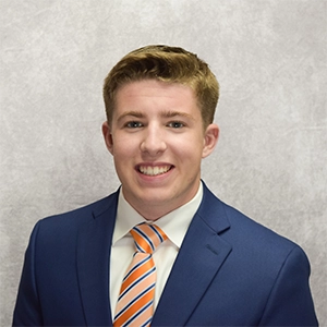 Headshot of a young professional man in a navy suit with a white shirt and orange-striped tie, smiling against a light gray background.