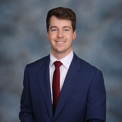 Professional headshot of a young man in a blue suit with a red tie, smiling against a soft gray background.