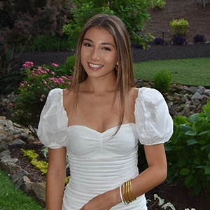 A young woman with long hair wearing a white puff-sleeve dress, smiling in a garden with colorful flowers.