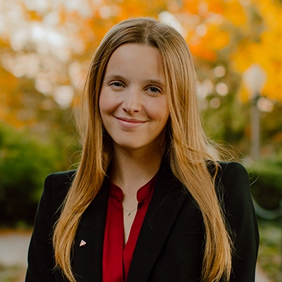 A young woman with long hair smiles confidently while wearing a black blazer over a red blouse, set against a backdrop of autumn foliage.