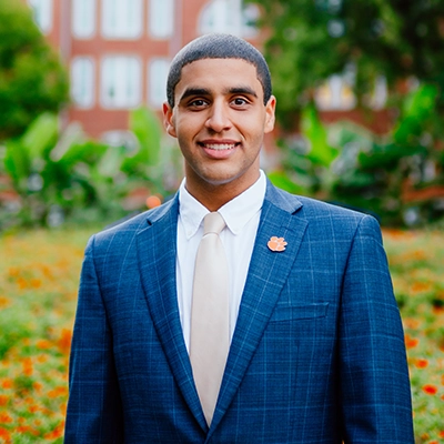 A young man in a suit stands smiling in a garden, featuring vibrant flowers and greenery in the background. He wears a small Clemson University logo on his lapel.