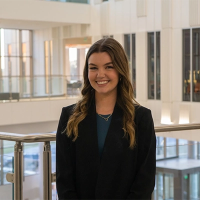 Professional headshot of a smiling woman in a blazer, standing in a modern, well-lit interior space.