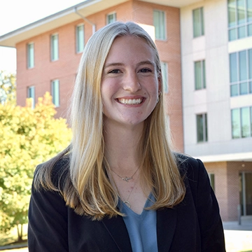 A smiling young woman with long blonde hair, wearing a black blazer, stands outdoors in front of a modern building.